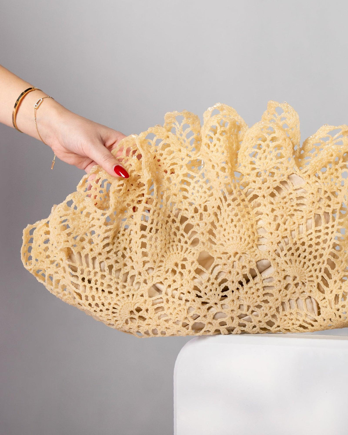 ALISADUDAJ Çantro beige crochet-lace clutch bag in polyester resin, close-up held by hand on a white stand, showing scalloped open-work edge