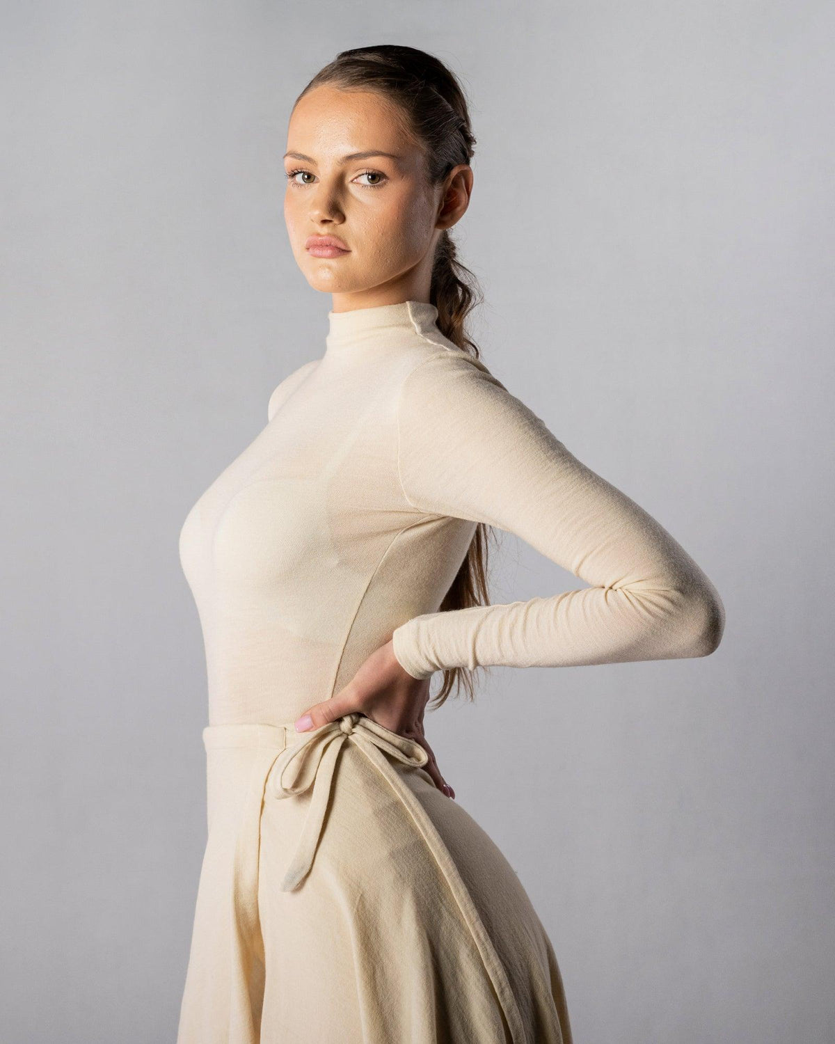 ALISADUDAJ CRAFT 07 off-white wool turtleneck and wrap skirt set, side three-quarter view with hand at the waist tie